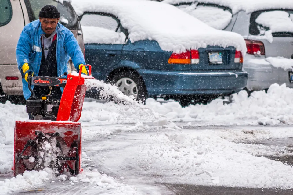 A man pushes a snow blower to clear the ground of an outdoor parking lot filled with cars.
